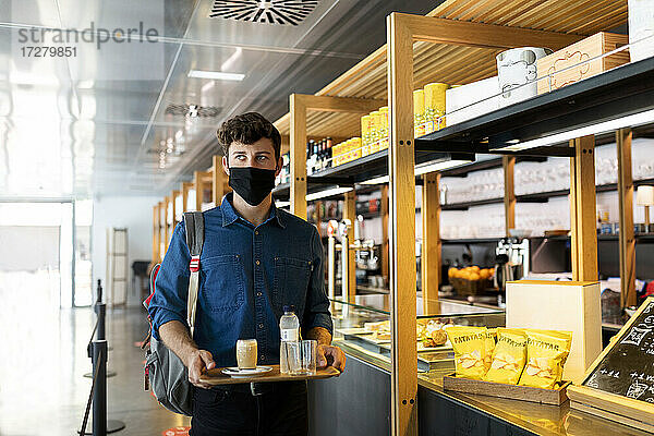 Geschäftsmann mit Gesichtsschutzmaske hält ein Tablett mit Getränken in einem Café während COVID-19
