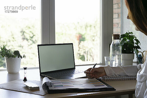 Arzt schreibt in Buch  während er zu Hause am Laptop arbeitet