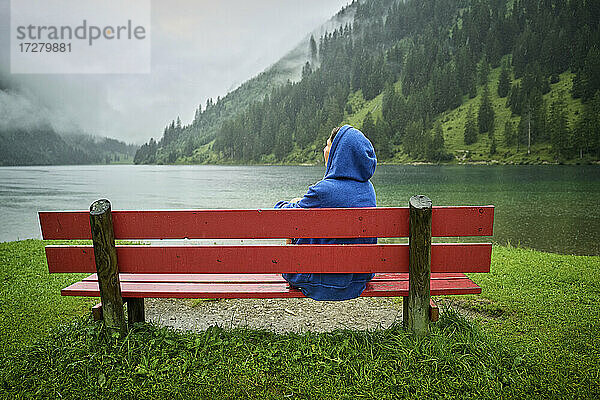 Frau mit blauer Kapuze sitzt auf einer Bank am Seeufer und schaut auf die Berge