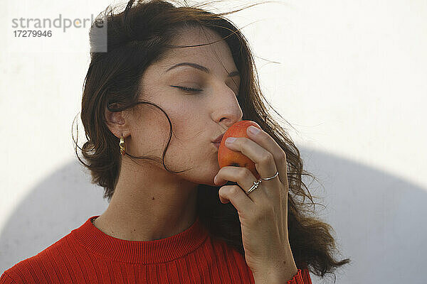 Junge Frau küsst einen Apfel  während sie an einem sonnigen Tag an der Wand steht