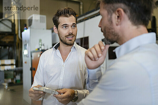Junger Geschäftsmann mit Metallgegenstand im Gespräch mit einem Kollegen in einer modernen Fabrik