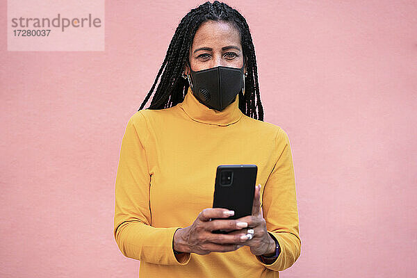 Ältere Frau mit Gesichtsmaske  die ein Mobiltelefon benutzt  während sie an einer rosa Wand steht