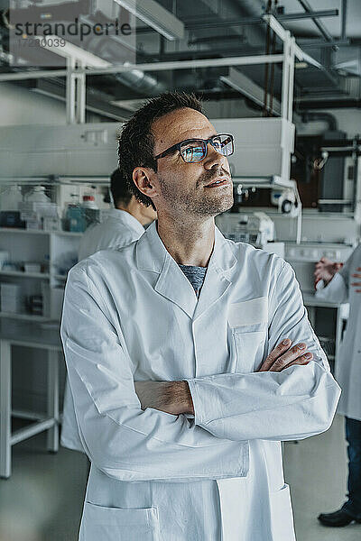 Männlicher Wissenschaftler mit verschränkten Armen  der wegschaut  während er mit einem Mitarbeiter im Hintergrund im Labor steht
