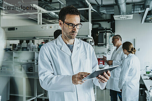 Wissenschaftlerin mit Brille  die ein digitales Tablet benutzt  während sie mit einem Mitarbeiter im Hintergrund in einem Labor steht