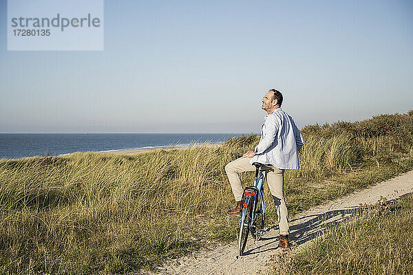 Lächelnder Mann  der die Aussicht betrachtet  während er mit dem Fahrrad am Strand vor dem klaren Himmel steht