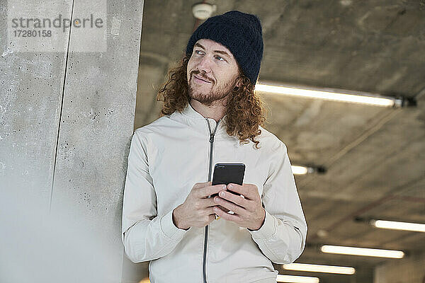 Lächelnder Hipster-Mann mit Strickmütze  der sein Smartphone benutzt  während er sich zu Hause an eine Säule lehnt