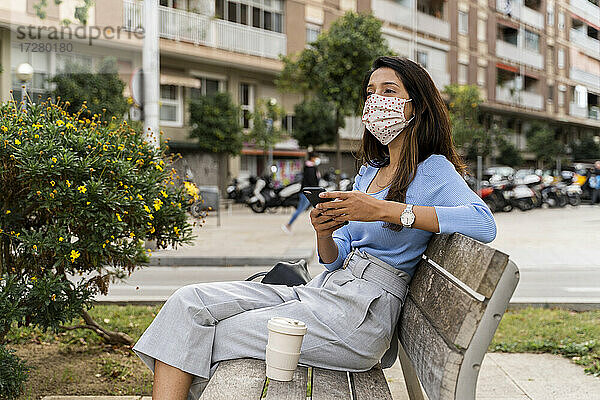Frau  die mit ihrem Smartphone wegschaut  während sie auf einer Bank in der Stadt sitzt  während COVID-19