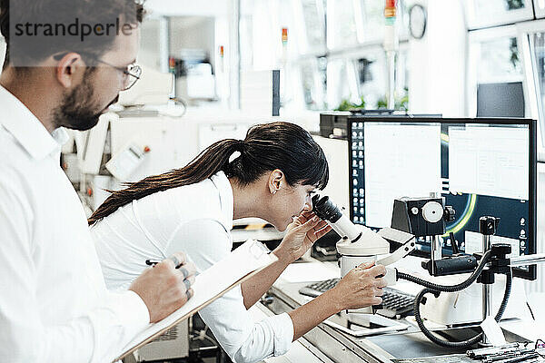 Weiblicher Geschäftsmann schaut durch ein Mikroskop  während er neben einem männlichen Kollegen im Labor steht