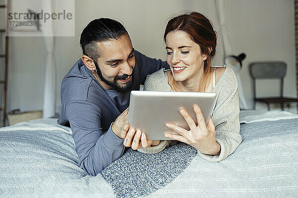 Lächelndes Paar  das ein digitales Tablet benutzt  während es zu Hause auf dem Bett liegt