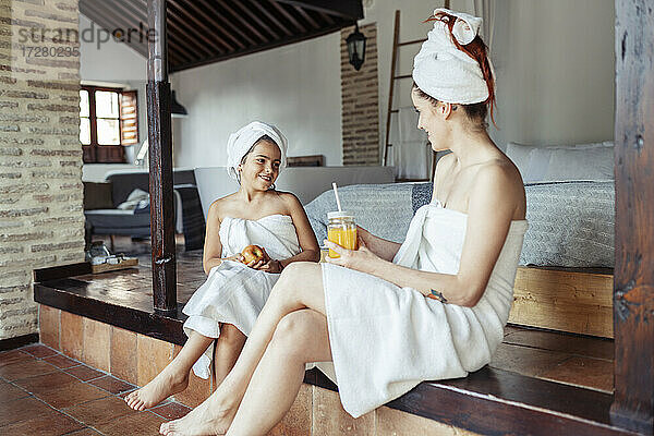Lächelnde Frau und Mädchen in Handtuch mit Obst und Saft beim Sitzen zu Hause
