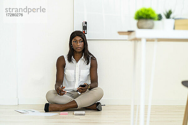 Geschäftsfrau  die ein Mobiltelefon benutzt  während sie im Büro auf dem Boden sitzt