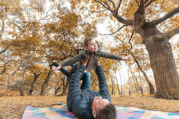 Junge  der sich vorstellt  dass er fliegt  gehalten von seinem Vater  auf einer Picknickdecke im Park im Herbst