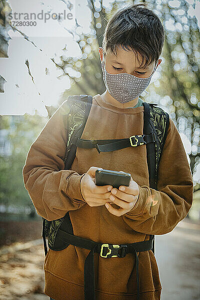 Junge  der ein Smartphone benutzt und eine Gesichtsmaske trägt  während er in einem öffentlichen Park steht