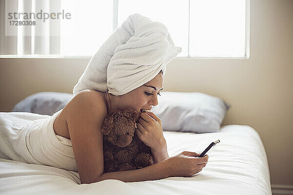 Junge Frau  eingewickelt in ein Handtuch  benutzt ein Smartphone  während sie zu Hause auf dem Bett liegt