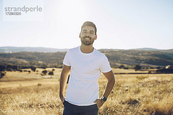 Lächelnder junger Mann steht mit den Händen in den Taschen auf einem Feld gegen den Himmel an einem sonnigen Tag