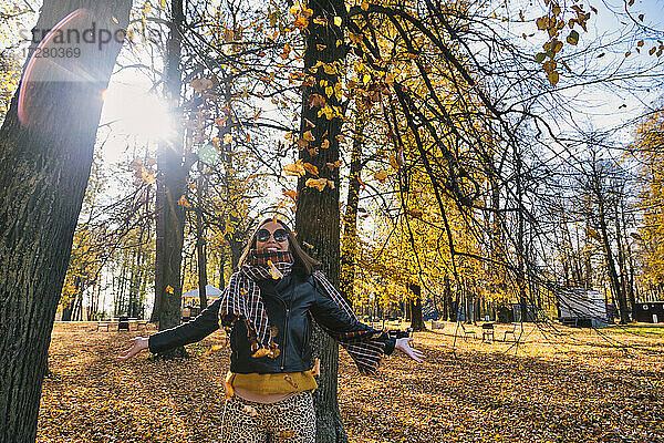 Glückliche Frau steht mit ausgestreckten Armen unter fallenden trockenen Herbstblättern vor Bäumen im Park