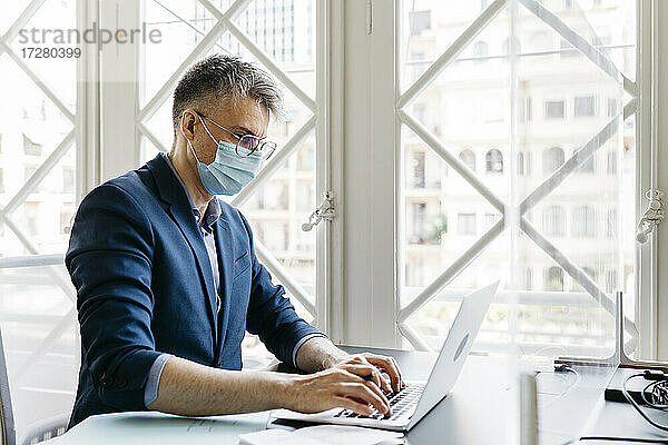 Männlicher Unternehmer mit Schutzmaske bei der Arbeit am Laptop vor dem Fenster im Büro