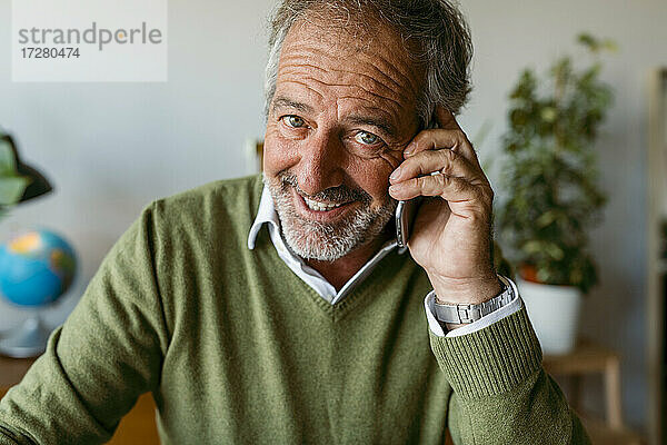 Lächelnder reifer Mann  der zu Hause sitzt und mit seinem Handy spricht