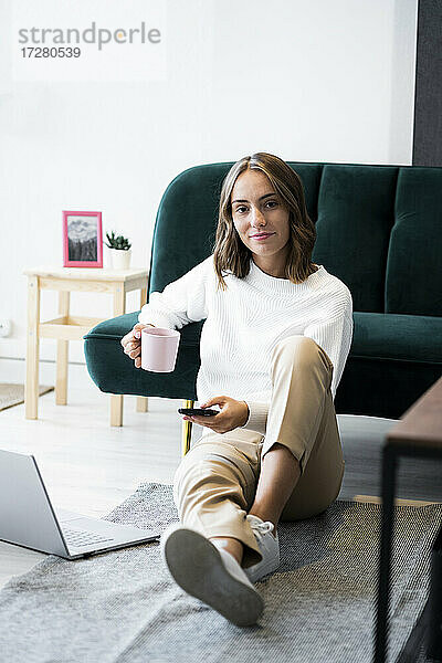 Lächelnde Geschäftsfrau mit Kaffeetasse und Mobiltelefon  während sie im Büro auf dem Boden sitzt