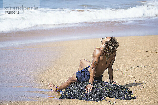 Junger Mann ohne Hemd entspannt sich auf einem Felsen am Strand