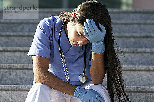 Junger Arzt mit Schutzhandschuh sitzt mit dem Kopf in der Hand auf einer Treppe
