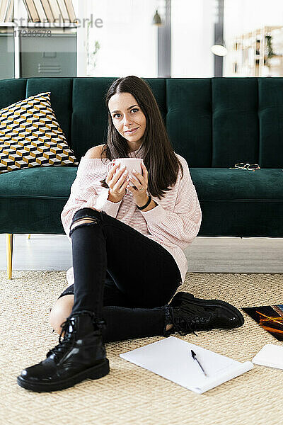 Schöne junge Frau sitzt mit Kaffeetasse und Notizblöcken auf Teppich vor Sofa zu Hause