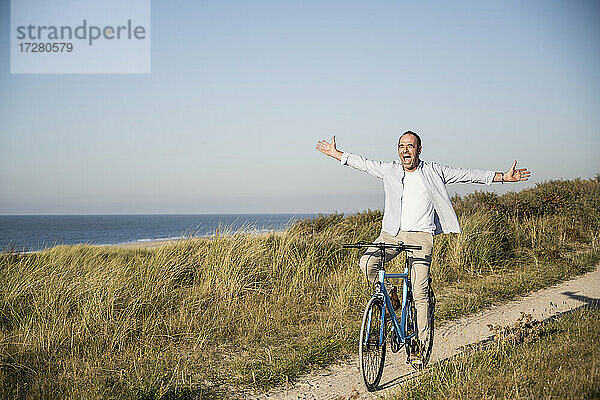 Glücklicher reifer Mann mit ausgestreckten Armen beim Fahrradfahren am Strand gegen den klaren Himmel