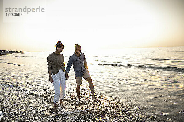 Glückliches junges Paar hält sich an den Händen  während es bei Sonnenuntergang am Strand spazieren geht