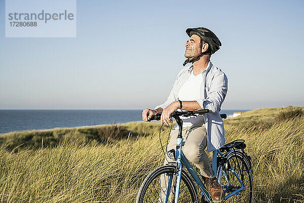 Lächelnder Mann mit geschlossenen Augen auf dem Fahrrad sitzend am Strand gegen den klaren Himmel