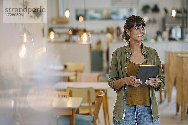 Lächelnde Unternehmerin  die wegschaut  während sie ein digitales Tablet in einem Café in der Hand hält