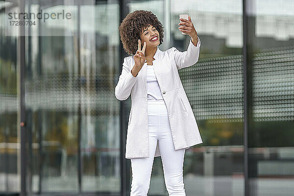 Lächelnde Geschäftsfrau  die eine Friedensgeste zeigt  während sie ein Selfie mit ihrem Mobiltelefon im Freien macht