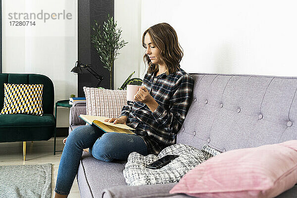 Junge Frau mit Kaffeetasse  die zu Hause auf dem Sofa sitzt und ein Buch liest