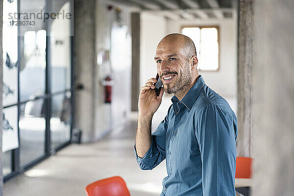Lächelnder Geschäftsmann  der auf einem Tisch im Büro sitzt und mit seinem Handy spricht