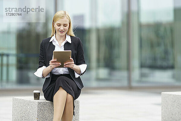 Blonde Geschäftsfrau benutzt ein digitales Tablet  während sie auf einer Bank vor einem Gebäude sitzt