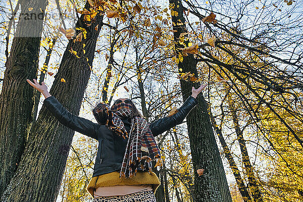 Frau wirft trockene Herbstblätter  während sie mit ausgestreckten Armen gegen Bäume im Park steht