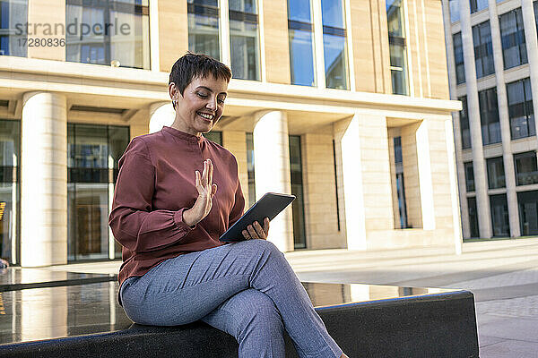 Frau winkt mit der Hand zu einem Videoanruf auf einem digitalen Tablet  während sie auf einer Bank vor einem Gebäude sitzt