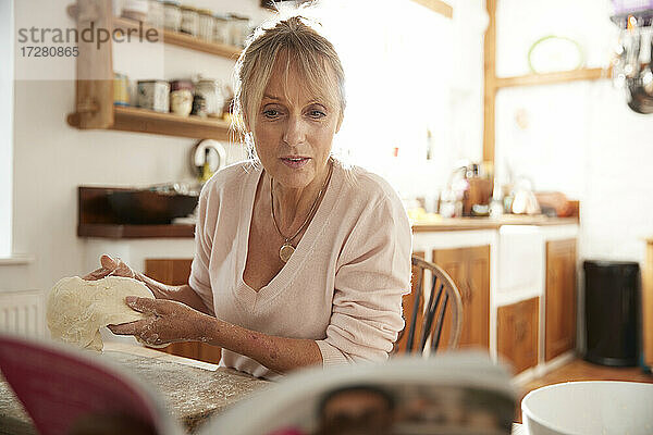 Ältere Frau knetet  während sie in der Küche sitzend ein Rezeptbuch liest