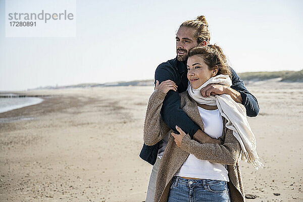 Lächelndes junges Paar schaut weg  während es sich am Strand gegen den klaren Himmel umarmt