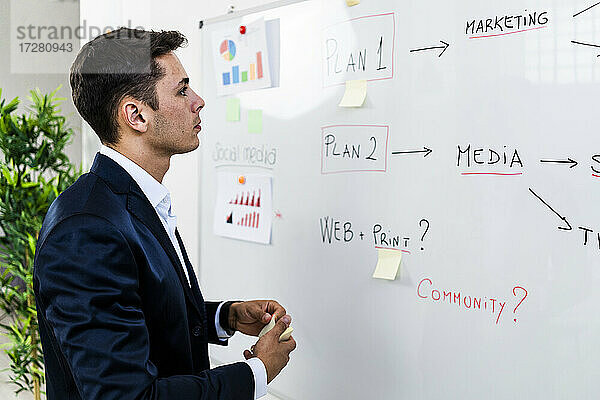 Männlicher Fachmann  der einen Geschäftsplan erstellt  während er eine Strategie auf einem Whiteboard am Arbeitsplatz betrachtet