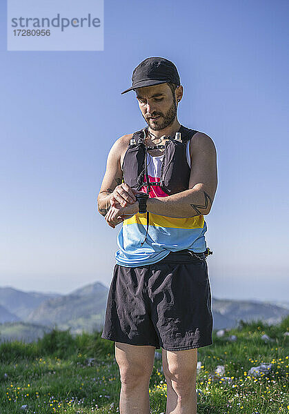 Mittlerer erwachsener Mann mit Mütze und Flaschengurt  der auf einem Berg stehend die Zeit auf der Uhr überprüft