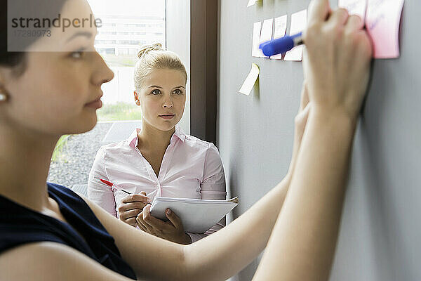Geschäftsfrau schreibt auf einen Klebezettel an der Wand  während sie mit einem Kollegen im Büro diskutiert