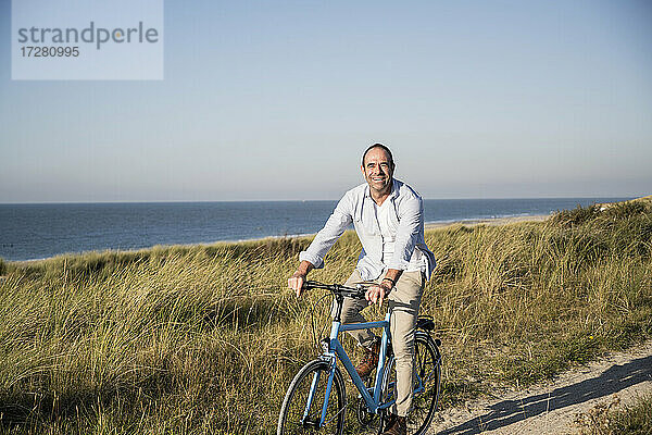 Lächelnder reifer Mann fährt Fahrrad am Strand gegen den klaren Himmel