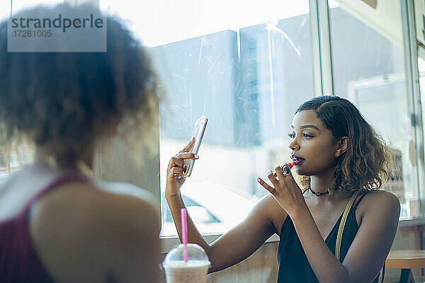 Junge Frau  die ein Mobiltelefon benutzt  um Lippenstift aufzutragen  während sie mit einer Freundin im Café sitzt