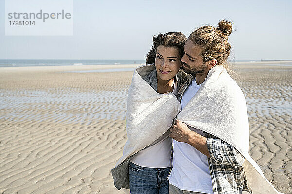 Romantisches junges Paar in Decke gehüllt am Strand stehend
