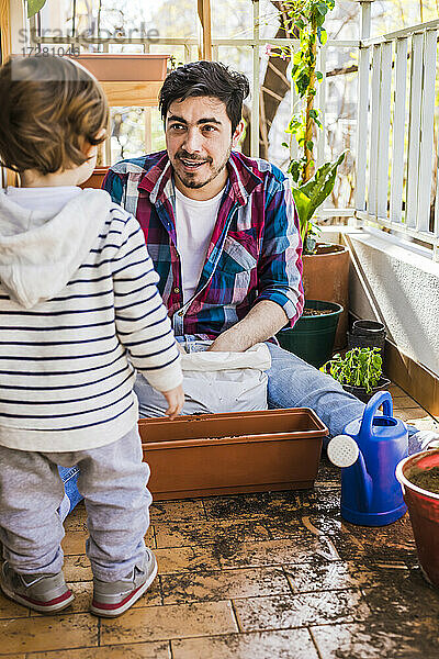 Mann schaut Junge an  während er eine Pflanze auf dem Balkon pflanzt