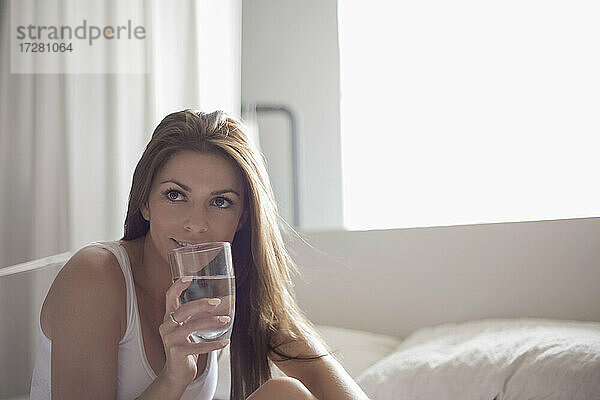 Junge Frau trinkt Wasser aus einem Glas im Schlafzimmer zu Hause