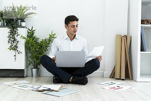 Gutaussehender männlicher Unternehmer  der Dokumente analysiert  während er mit seinem Laptop auf dem Boden vor einer weißen Wand im Büro sitzt