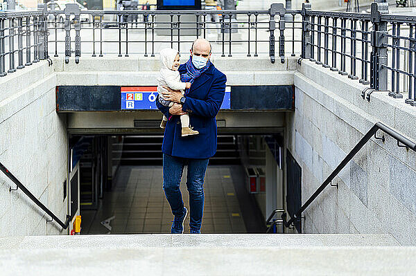 Mann mit Gesichtsmaske trägt Baby beim Gehen auf einer Treppe in der Stadt