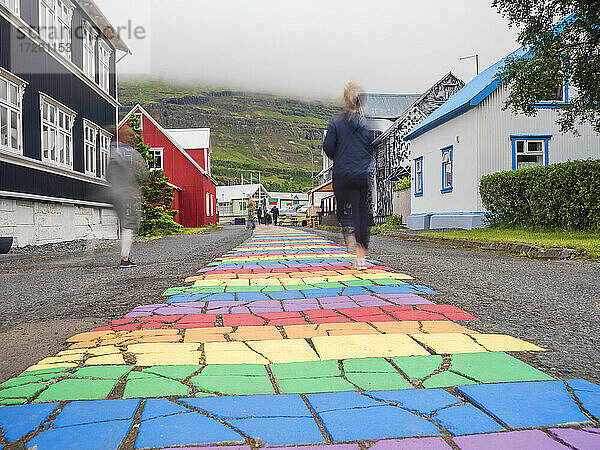 Island  Seydisfjordur  Menschen gehen entlang der regenbogenfarbenen Asphaltstraße inmitten einer abgelegenen Stadt