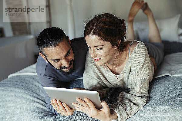 Mann und Frau benutzen ein digitales Tablet  während sie zu Hause auf dem Bett liegen